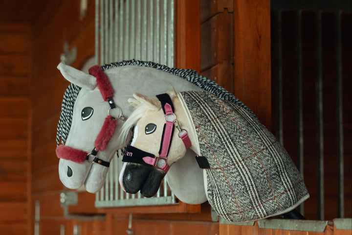Dva plyšoví hobby horse s uzdečkami a dečkami odpočívají na dřevěné přepážce stáje a připomínají skutečné koně v boxu. Ideální pro fanoušky Hobby Horsing, bílý a šedý kůň mají propracované detaily a výstroj.