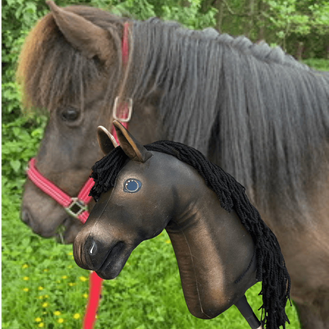 Hobby Horse Koník podle fotografie od Hobby Horse LarDen vedle skutečného hnědého koně s červenou ohlávkou; hnědý koník na tyči s černou přízovou hřívou, v exteriéru se zelení a stromy.