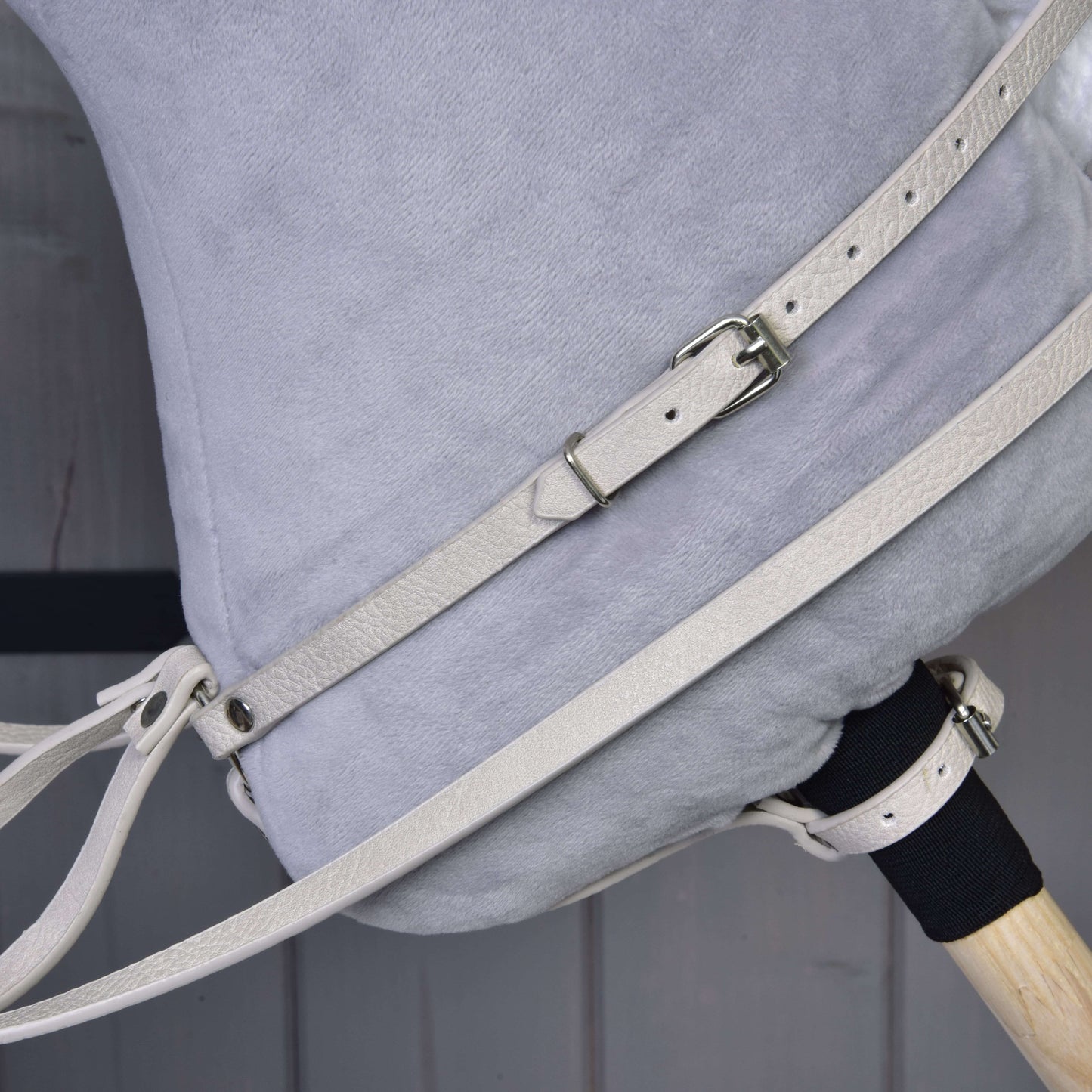 Detail boku plyšového Hobby Horse od Hobby Horse LarDen se světle šedou látkou a koženkovým setem Aqua – uzdečka, otěže a martingal – s přezkami, plus část dřevěné rukojeti před dřevěným pozadím.