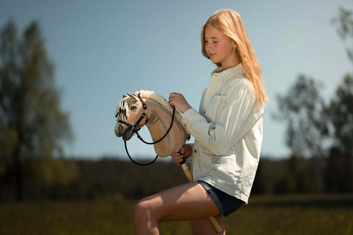 Mladý člověk s dlouhými blond vlasy, ve světlé bundě a kraťasech, si užívá Hobby Horsing na hobby horse venku na slunné travnaté louce se stromy v pozadí.