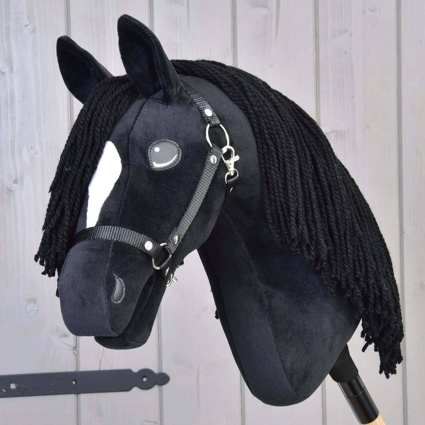 Hobby Horse Cloony s černou ohlávkou od Hobby Horse LarDen, plyšový hřebec American Quarter Horse z černé látky s bílou skvrnou na hlavě, knoflíkovýma očima, černou přízovou hřívou a uzdečkou na světlém dřevěném pozadí.