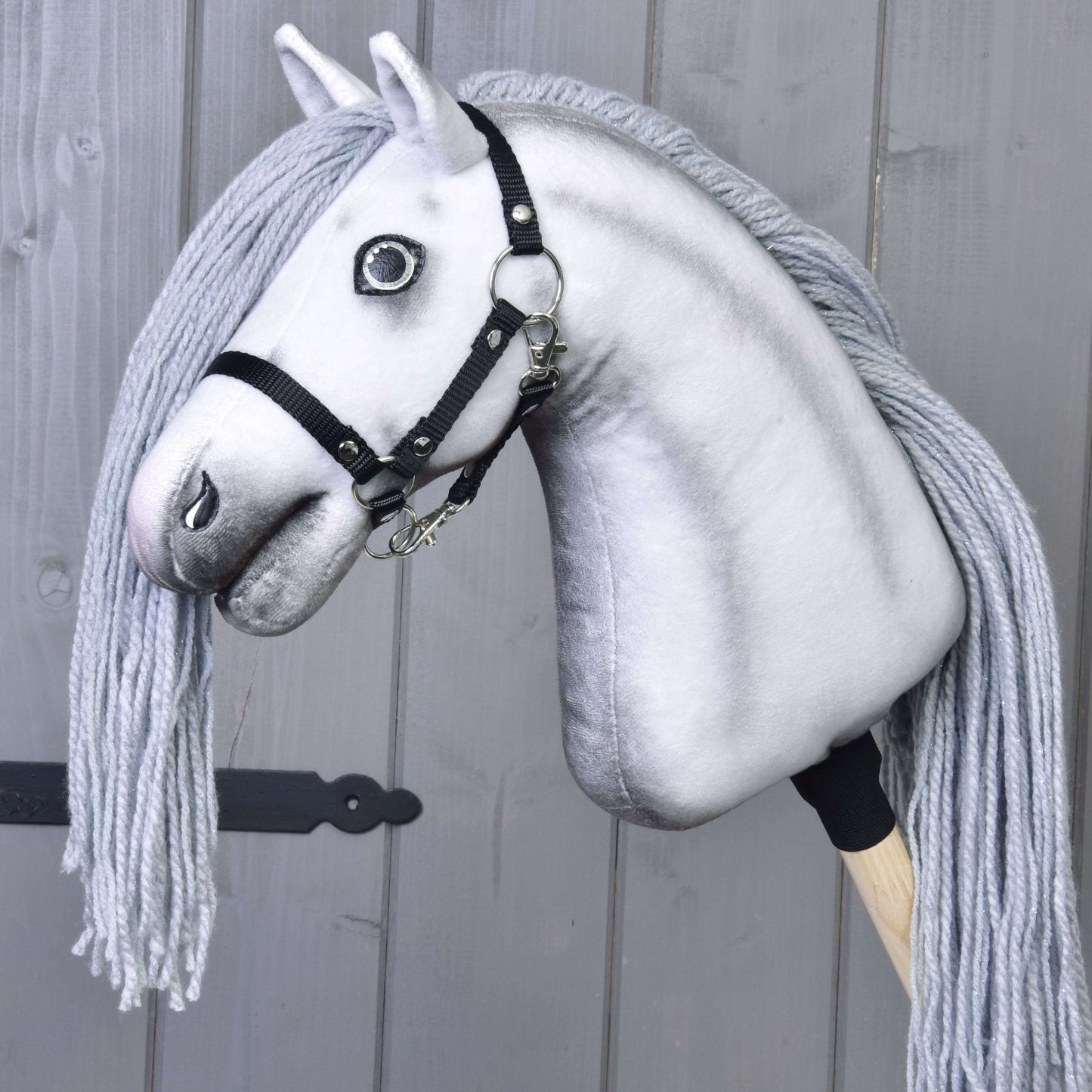 Hobby Horse jednorožec Silver s uzdou a ohlávkou od Hobby Horse LarDen s černou uzdou, ohlávkou a dlouhou hřívou z příze, připomínající kouzelného jednorožce. Zobrazeno na pozadí šedé dřevěné stěny.