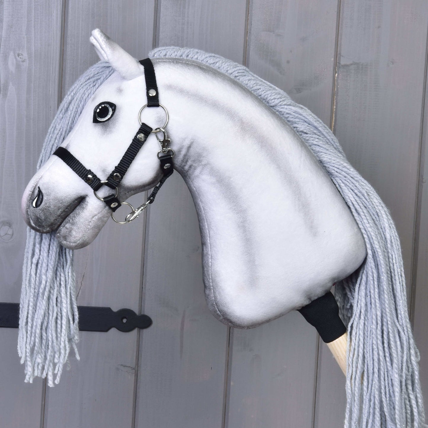 Hobby Horse jednorožec Silver s uzdou a ohlávkou od Hobby Horse LarDen s bílou hlavou, černou uzdou a ohlávkou, černýma knoflíkovýma očima a dlouhou šedou hřívou, zobrazený na šedém dřevěném pozadí.