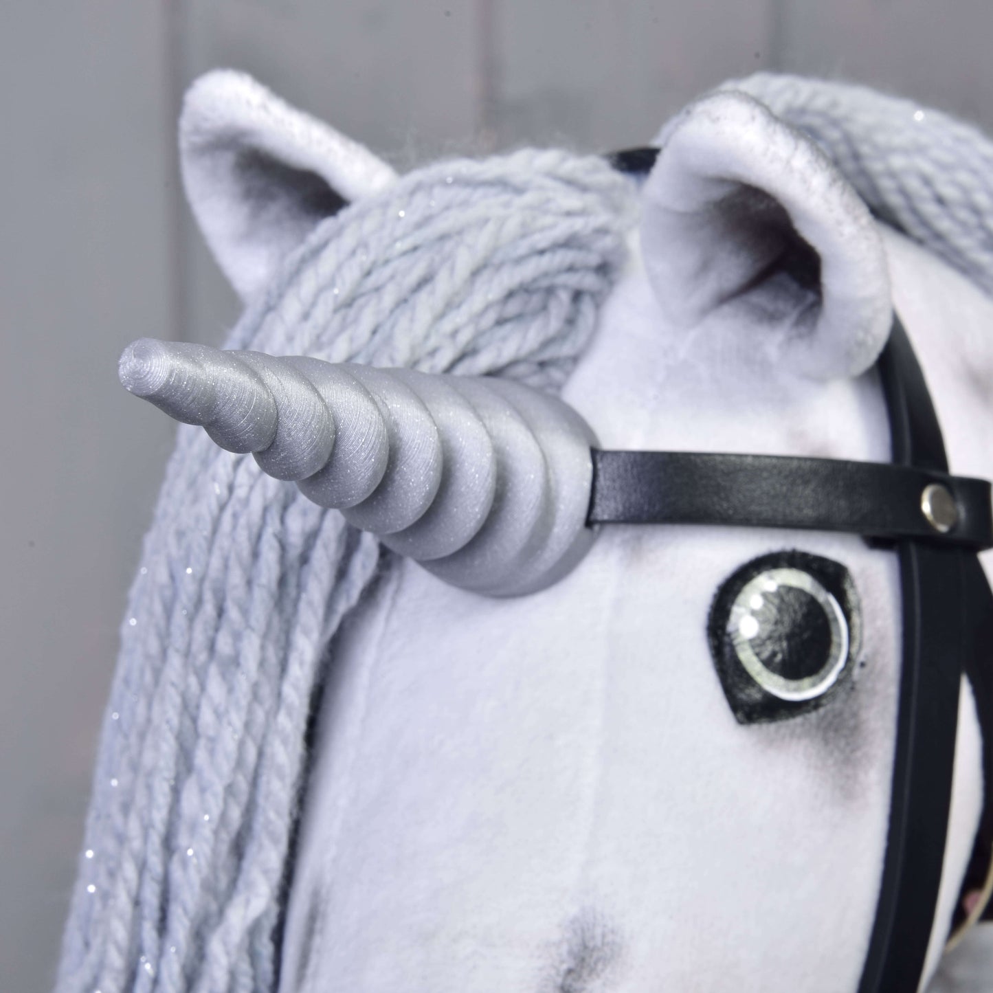 Detail Hobby Horse jednorožec Silver s uzdou a ohlávkou od Hobby Horse LarDen se šedým spirálovým rohem, knoflíkovým okem, dlouhou šedou hřívou z příze a černou uzdou na neutrálním pozadí.