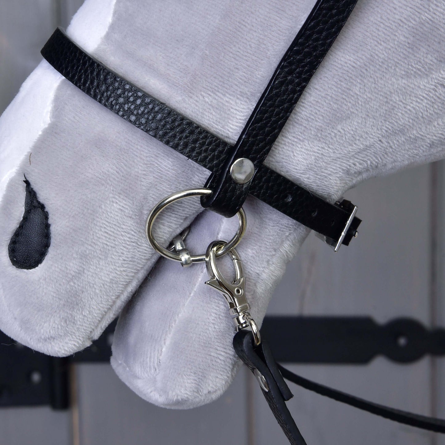 Detail hlavy bílého plyšového koně s Kompletní černou uzdečkou s kamínky od Hobby Horse LarDen, s odnímatelnými otěžemi a anglickým nánosníkem s kovovým udidlem, na dřevěném pozadí s viditelným tmavým kováním.