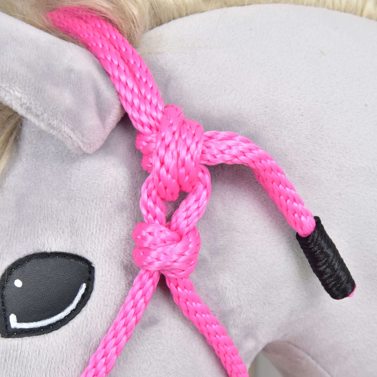 Detail hlavy plyšového koně s Hobby Horse LarDen Parelka Pink, uvázanou kolem ucha. Kůň má světle šedé tělo, malované černé oko s bílými odlesky a světle blond hřívu.