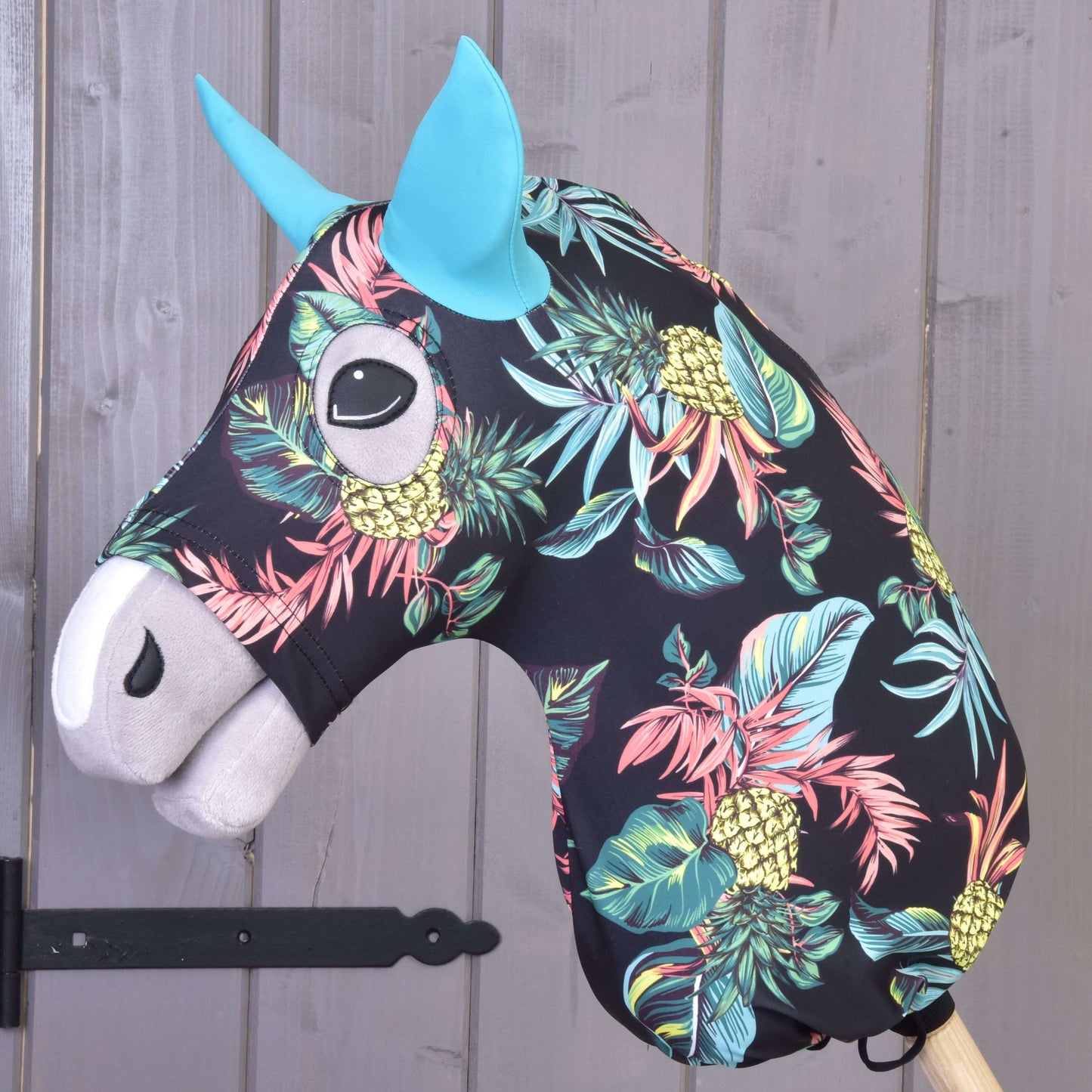 Hobby Horse ochranný oblek Pineapple (Lycra) od Hobby Horse LarDen má černou hlavu s výrazným potiskem ananasů a listů, modré uši a bílý čenich, je nasazený na dřevěné tyči a zobrazený před šedou dřevěnou stěnou.