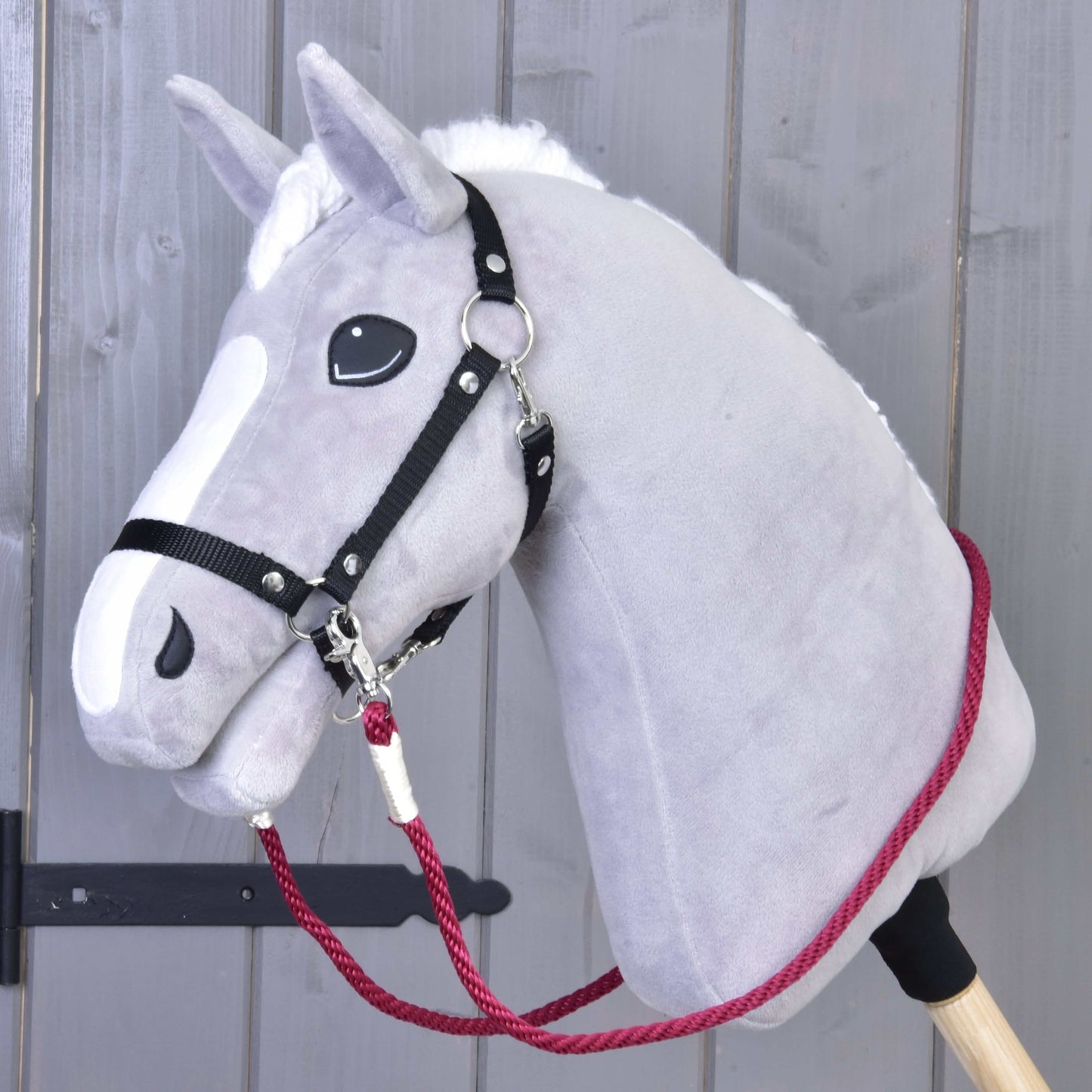 Otěže Czi-Czi od Hobby Horse LarDen na šedém plyšovém Hobby Horse s bílou hřívou, černou uzdečkou a ručně vyráběnými červenými otěžemi, nasazeném na dřevěné tyči před šedou dřevěnou stěnou.