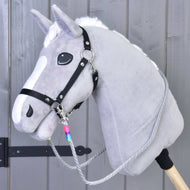 Hobby Horse LarDen Otěže Manhattan, šedý Hobby Horse s bílou hřívou, černou ohlávkou, ručně vyráběnými otěžemi a dřevěnou tyčí, zobrazený před šedou dřevěnou stěnou.
