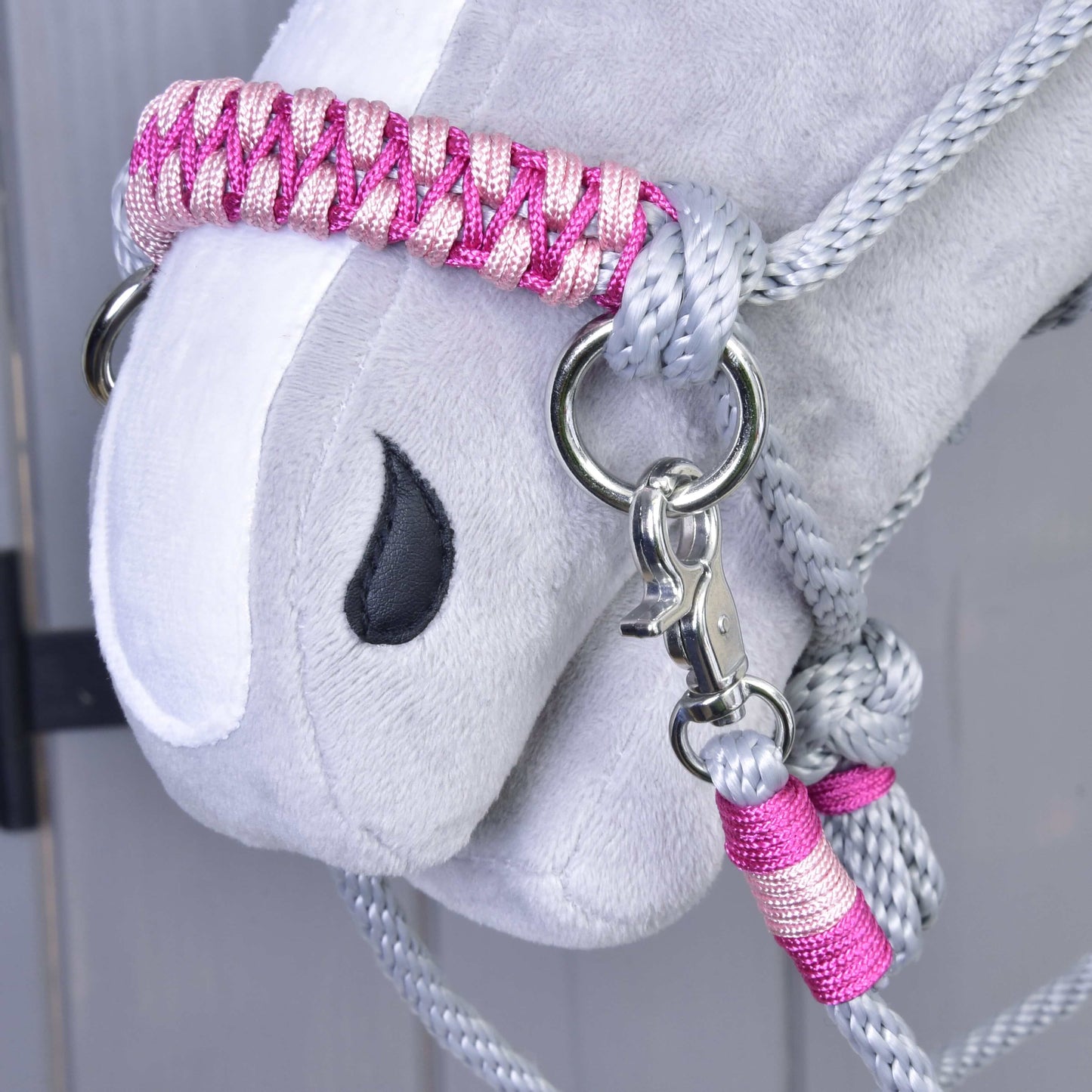 Detail šedé plyšové hlavy Hobby Horse LarDen s Provazovým setem Rosie: růžovo-stříbrná pletená ohlávka a vodítko, černé vyšívané oko a kovová karabina na světlém dřevěném pozadí.