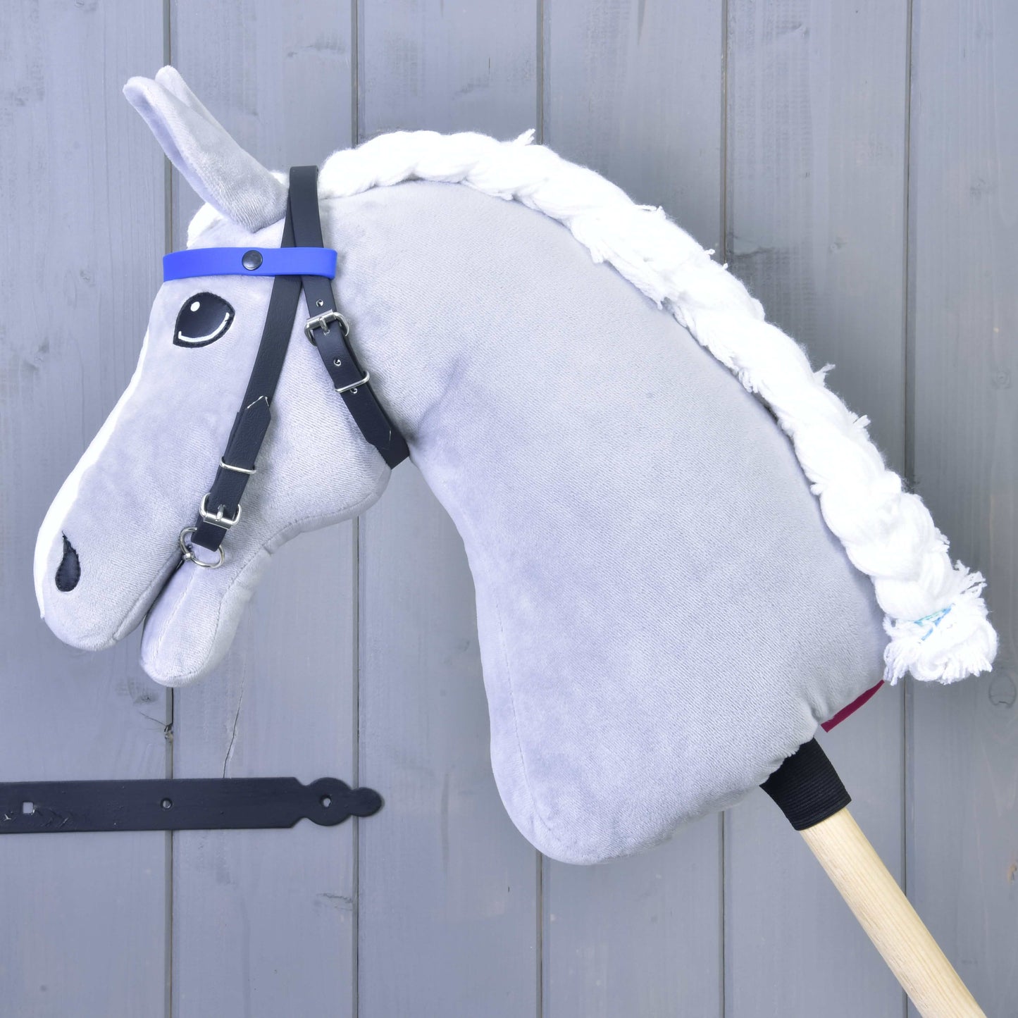 Uzdečka bez nánosníku modrá se 2 čelenkami (vel. M) od Hobby Horse LarDen na plyšovém Hobby Horse s bílou přízovou hřívou, připevněném na dřevěné tyči a opřeném o šedou dřevěnou stěnu.