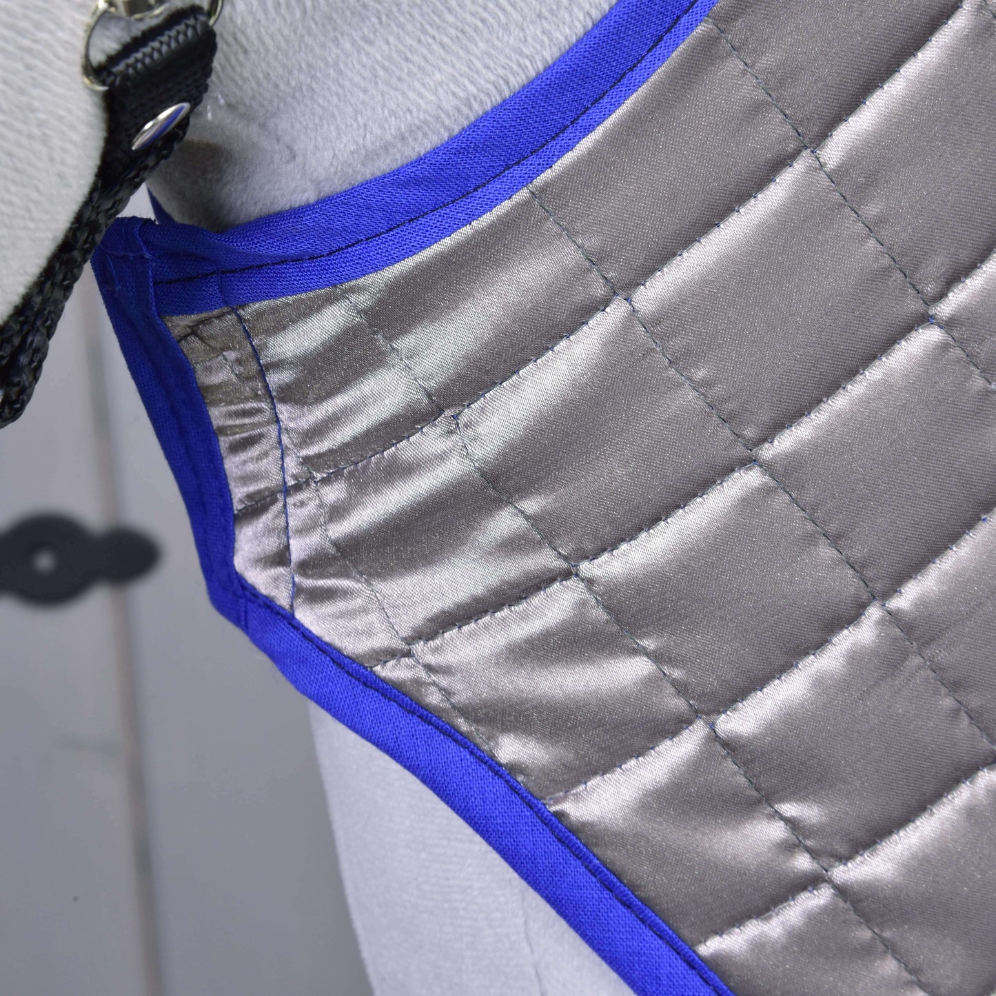 Detail Deka DeLuxe Blue oboustranná od Hobby Horse LarDen, prošívané stříbrno-šedé tréninkové deky pro koně s modrým lemováním a černým popruhem, přehozené přes světle zbarveného koně pro dodatečnou ochranu.