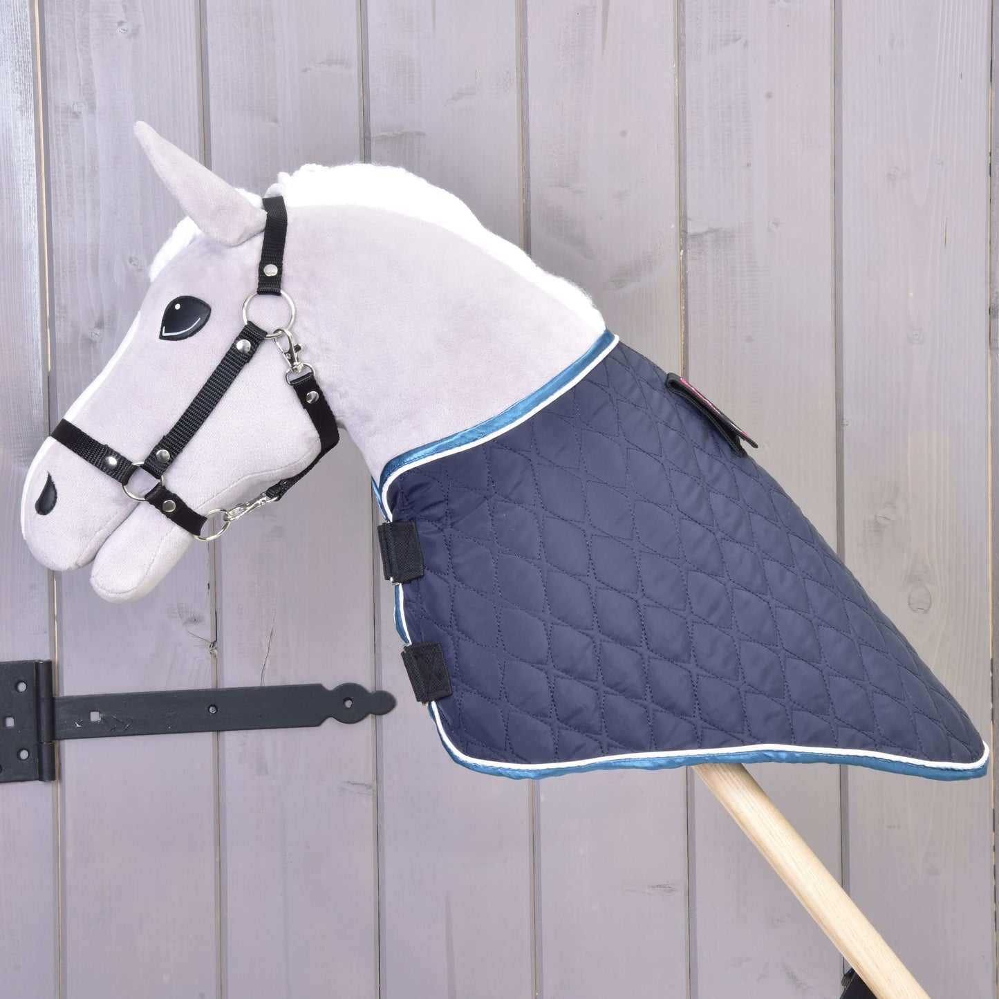 Plyšový Hobby Horse LarDen s bílou hřívou, s dekou s odepínatelným krkem Mandarin, opřený o světlou dřevěnou stěnu.