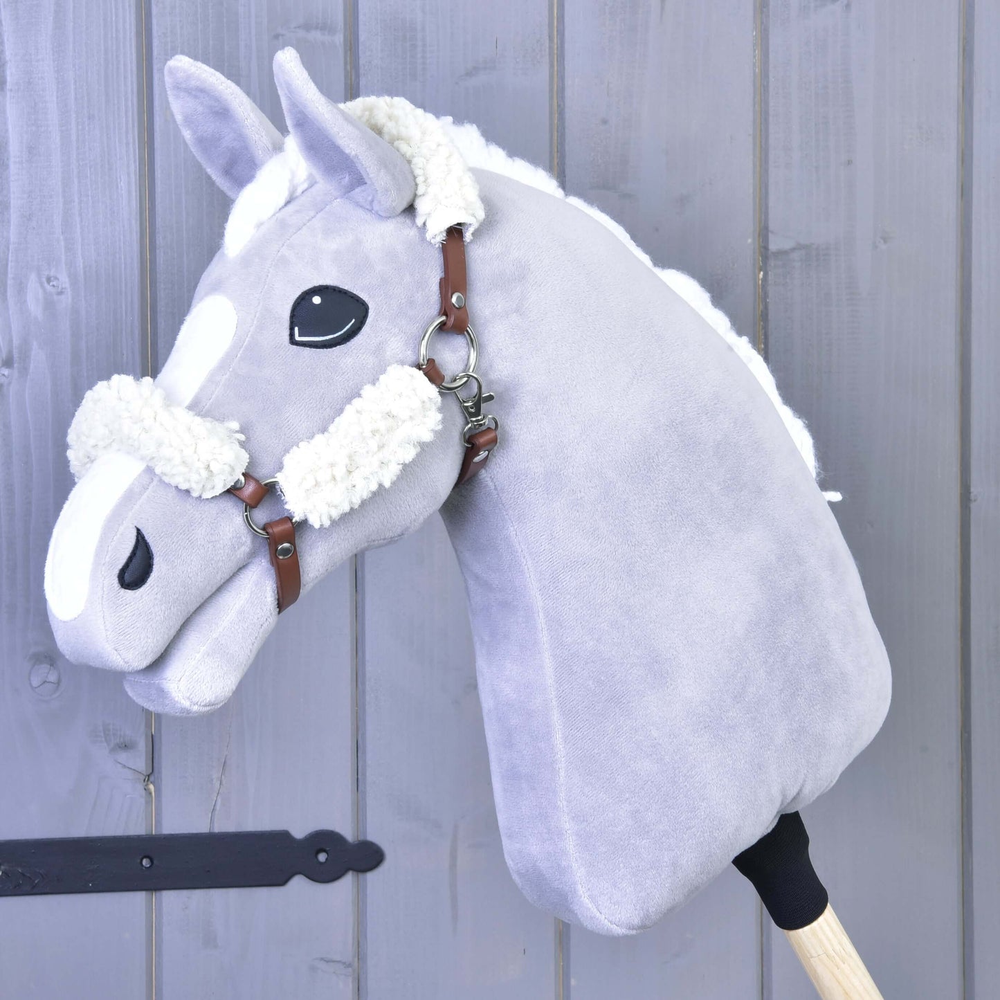 Hnědá ohlávka z koženky s kompletním beránkem od Hobby Horse LarDen na plyšovém světle šedém Hobby Horse s bílou hřívou a odznaky, nasazeném na dřevěné tyči a vyfotografovaném před pozadím z šedých dřevěných panelů.