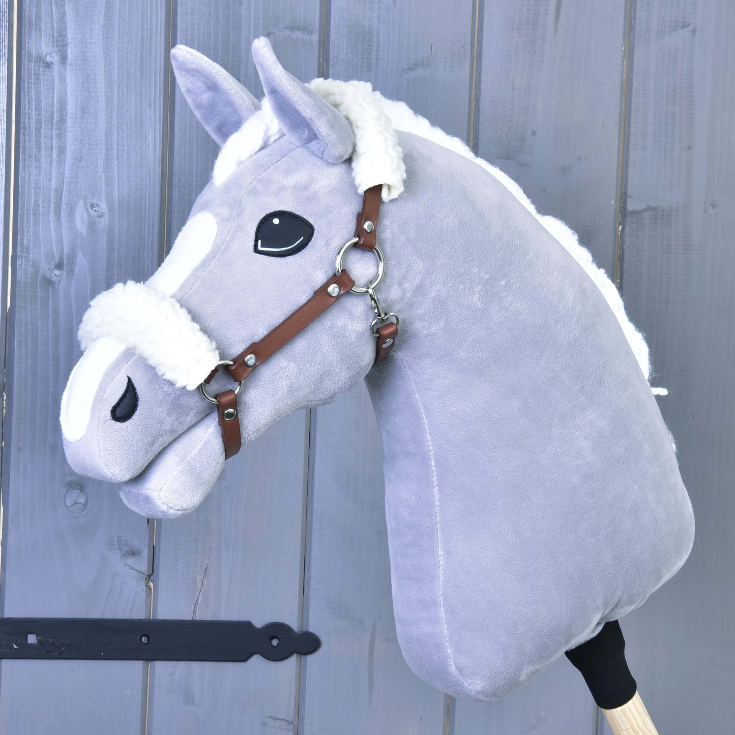 Plyšový šedý Hobby Horse od Hobby Horse LarDen s bílou hřívou a hnědou ohlávkou z koženky s beránkem, nasazený na dřevěné tyči na pozadí šedé dřevěné stěny.