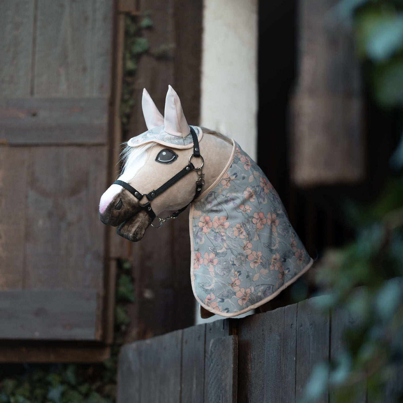 Realistický Hobby Horse, ideální pro Hobby Horsing, s uzdečkou a modrou dekou s květinovým vzorem je přehozený přes tmavý dřevěný plot, na pozadí jsou dřevěné okenice a svěží zeleň.
