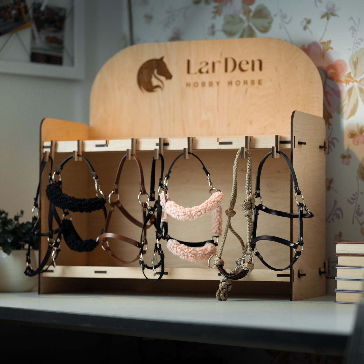Dřevěný stojan s nápisem LarDen Hobby Horse drží několik barevných uzdeček a ohlávek pro hobby horse – ideální pro fanoušky hobby horsing. Stojan je umístěný na bílém stole vedle hromady knih, na pozadí je květinová tapeta.