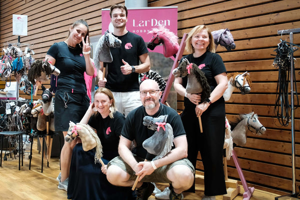Pět usměvavých dospělých pózuje uvnitř se svými hobby horse a slaví Hobby Horsing. Čtyři stojí a jeden klečí; všichni mají na sobě černá trička. Za nimi je dřevem obložená stěna a růžový nápis „LarDen“, s vystavenými různými hobby horse.
