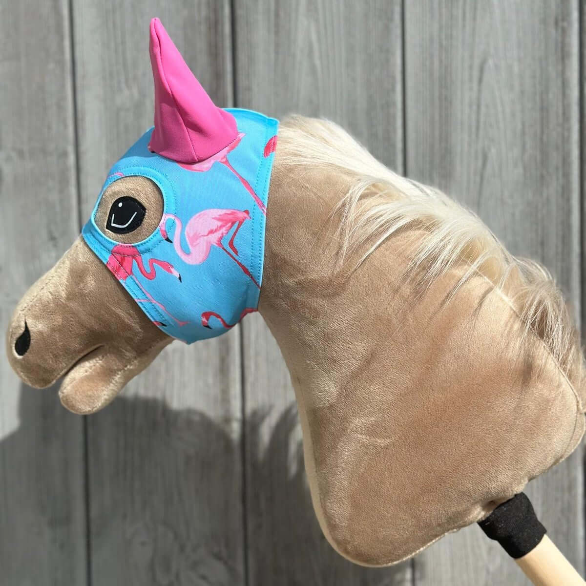 Plyšový Hobby Horse, ideální pro Hobby Horsing, má světle hnědou srst a bílou hřívu, modrý návlek na hlavu s růžovými plameňáky a velký růžový jednorožčí roh, na dřevěném pozadí.