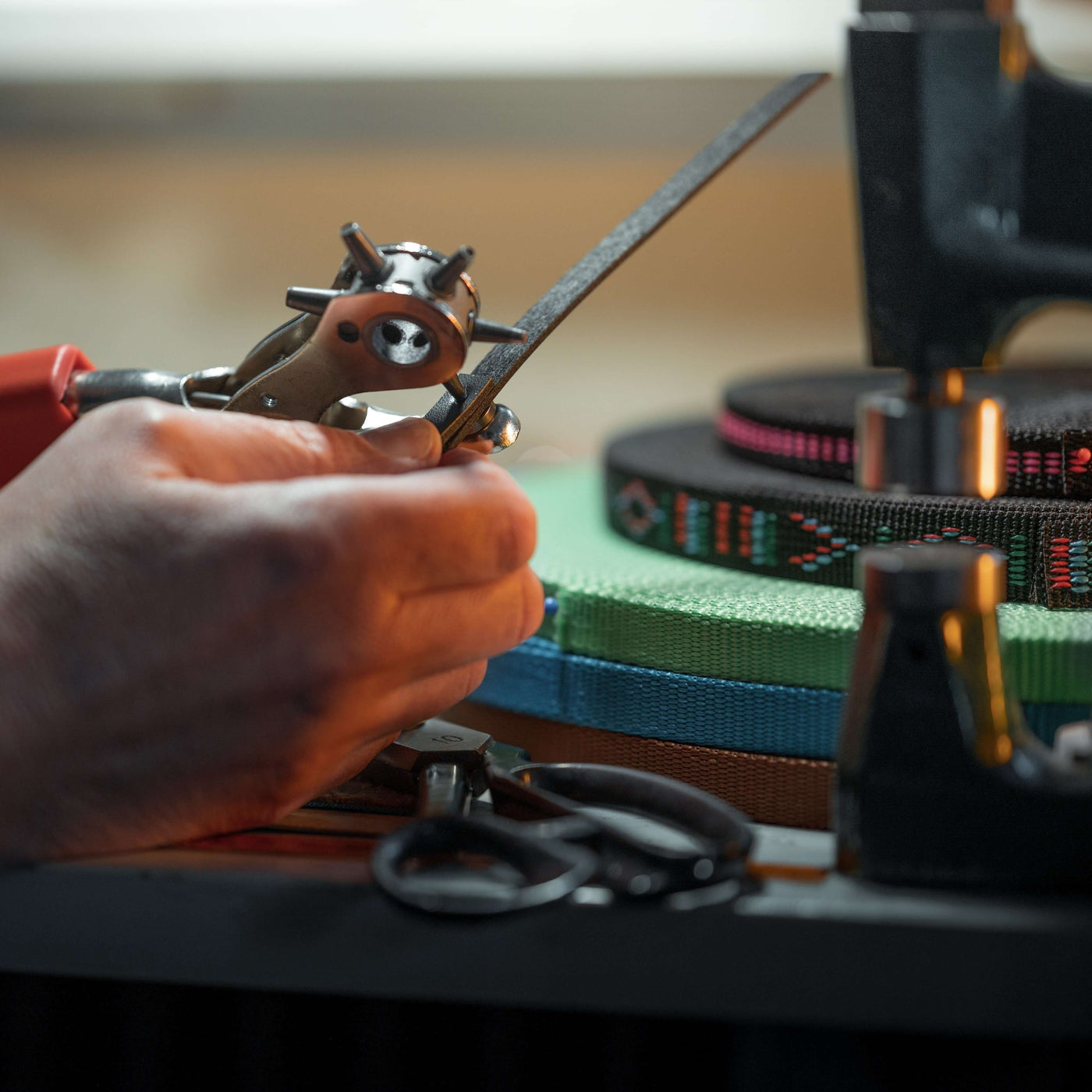 Detail rukou osoby používající děrovací nástroj na kůži – ideální materiály pro výrobu výstroje pro Hobby Horse – s rolemi barevných popruhů, šicími nástroji a nůžkami rozloženými na pracovním stole v pozadí.