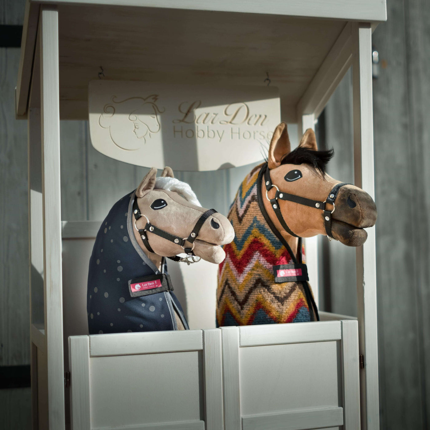 Dva plyšoví Hobby Horse s uzdečkami a dekami, jeden s modrým puntíkovaným potahem a druhý s barevným cikcak vzorem, jsou vystaveni vedle sebe v bílém dřevěném stánku označeném LarsDen Hobby Horsing.