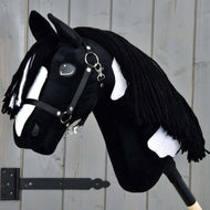 Hobby Horse Merlin s černou ohlávkou od Hobby Horse LarDen, inspirovaný hřebcem plemene Barock Pinto, má černobílé skvrny a zapletenou přízovou hřívu, upevněný před šedou dřevěnou stěnou.