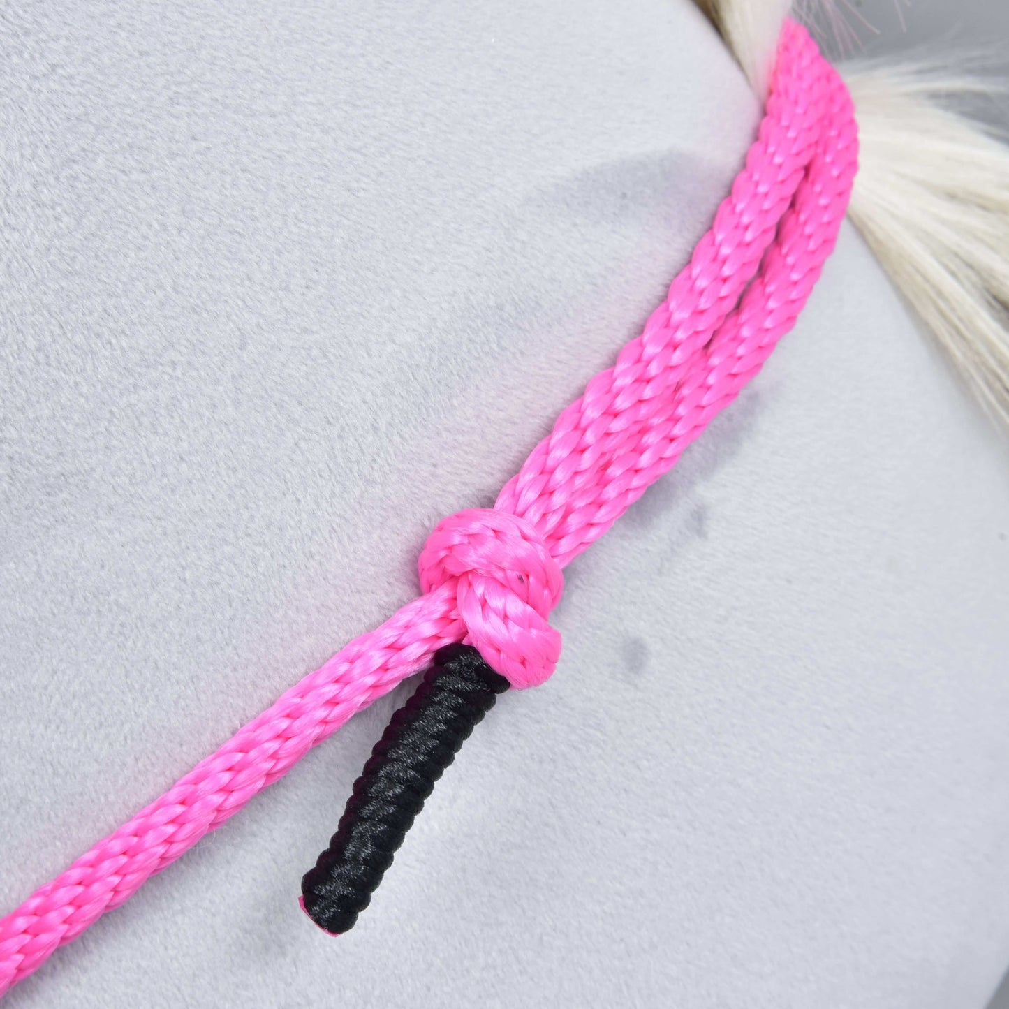 Detail Nákrčníku Pink Festive od Hobby Horse LarDen – výrazně růžové ručně vyrobené cordeo lano s pramenem s černým zakončením, uvázané do uzlu kolem světle šedého povrchu. Ideální pro ježdění bez uzdečky; pozadí je hladké s jemnými stopami.