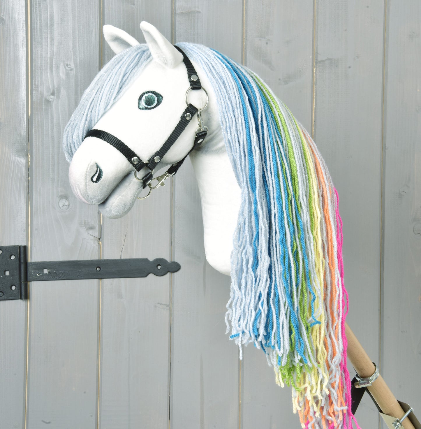 Hobby Horse Rainbow New od Hobby Horse LarDen má ručně vyrobený bílý obličej, modré oči, černou uzdečku a duhovou hřívu z modré, zelené, žluté a růžové příze na světle šedém dřevěném pozadí pro pohádkový dojem.