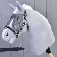 Plyšová šedá hlava Hobby Horse s bílou hřívou, s ohlávkou od Hobby Horse LarDen v černé barvě se šedými třásněmi a šedým nánosníkem, na dřevěné tyči, před dřevěnou stěnou.