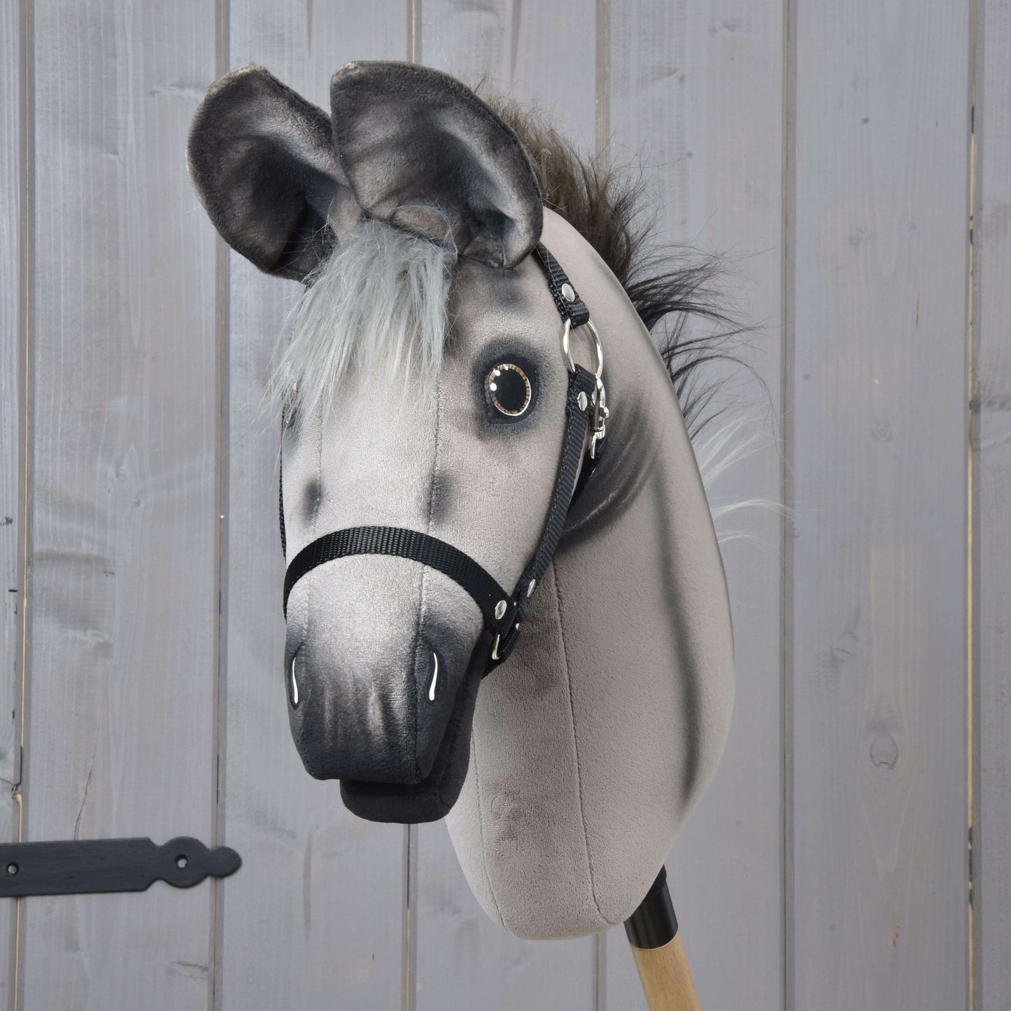 Hobby Horse osel Olin s černou ohlávkou od Hobby Horse LarDen, šedá oslí hračka na dřevěné tyči s malovanými detaily a černým postrojem, před světle šedým dřevěným obkladem.