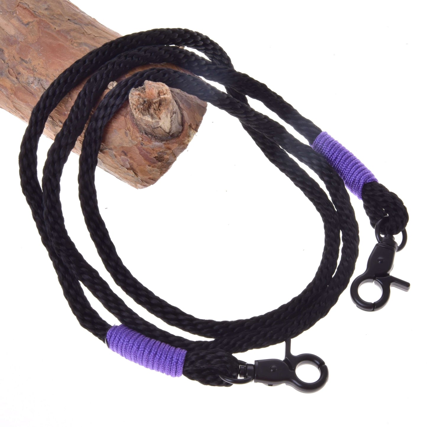 Otěže Basic Lila od Hobby Horse LarDen s černým pleteným polypropylenovým lanem, fialově omotanými konci a černými otočnými karabinami, ideální jako ručně vyráběné otěže nebo otěže pro Hobby Horse, elegantně stočené na dřevěném polenu.