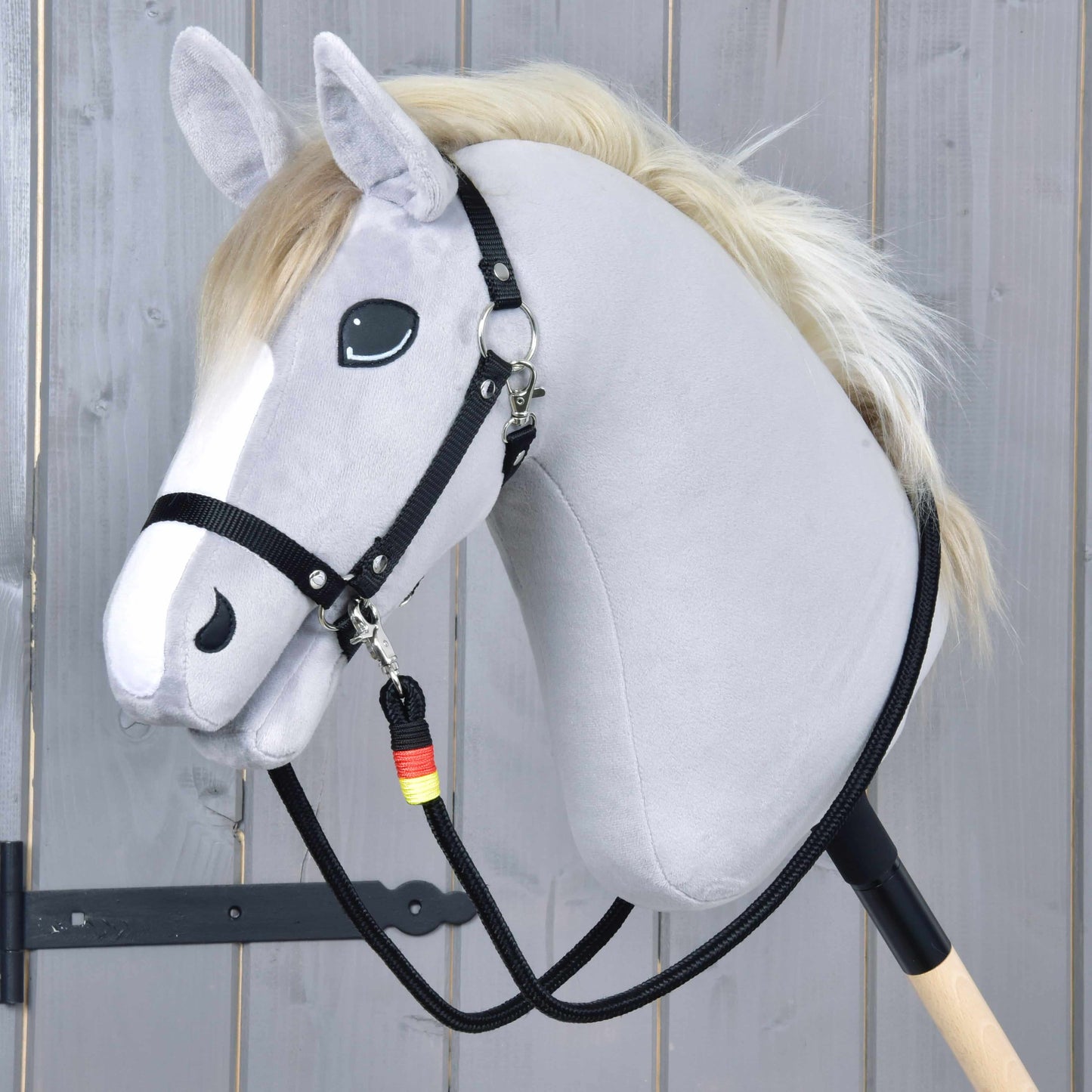 Otěže Emblem od Hobby Horse LarDen na šedém plyšovém Hobby Horse s bílou hřívou, černou uzdečkou a ručně vyráběnými polypropylenovými otěžemi na dřevěné tyči, na pozadí světlých dřevěných prken.