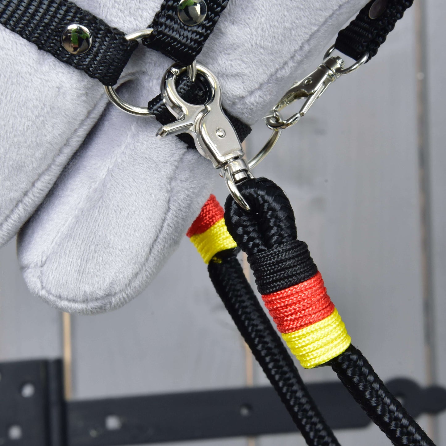 Detail otěží Otěže Emblem od Hobby Horse LarDen – černé otěže z polypropylenového lana s červenou a žlutou omotávkou, připnuté ke kovovému kroužku na černém řemínku, připevněné k šedé plyšové koňské hlavě na dřevěném pozadí.
