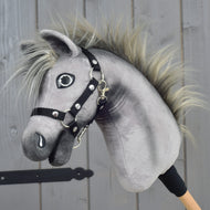 The Hobby Horse LarDen “Hobby Horse Prestige with black halter, size XS” features a gray plush design with a spiky mane and black halter, inspired by the elegance of an English Warmblood. It is mounted on a wooden stick and displayed against a wooden panel background.