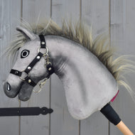 The Hobby Horse Prestige with black halter, size XS by Hobby Horse LarDen is a plush gray hobby horse head inspired by the elegance of an English Warmblood sport horse. It features an expressive embroidered face, a wild mane, and a secure black halter, mounted on a wooden stick handle. Posed against a wooden background, it’s perfect for imaginative show jumping adventures.