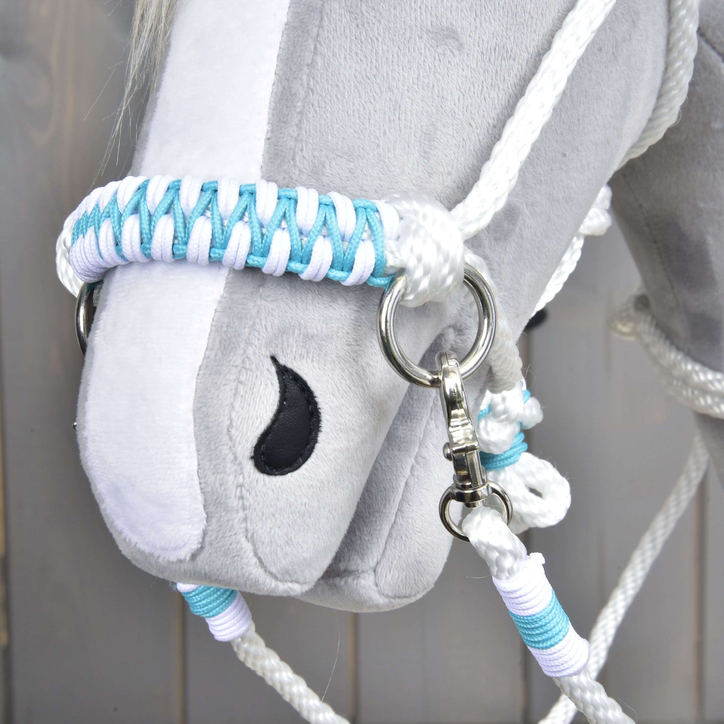 Detail šedé plyšové hlavy Hobby Horse LarDen s Provazovým setem White Queen – bílou, modrou a světle fialovou pletenou ohlávkou a otěžemi – s černě vyšitým okem a kovovým kroužkem na postroji.