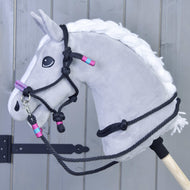 Provazový set Princess od Hobby Horse LarDen na šedém Hobby Horse s bílou hřívou, detailní černou ohlávkou a otěžemi a růžovými a tyrkysovými akcenty, nasazený na dřevěné tyči na pozadí šedých prken.