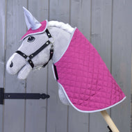 Set Hot Pink Silver od Hobby Horse LarDen: plyšový Hobby Horse se šedým obličejem, černou uzdečkou, bílou hřívou, prošívaným setem deky v sytě růžové a bílé barvě a ladící čabrakou na uši, vše na dřevěné tyči před dřevěnou stěnou.