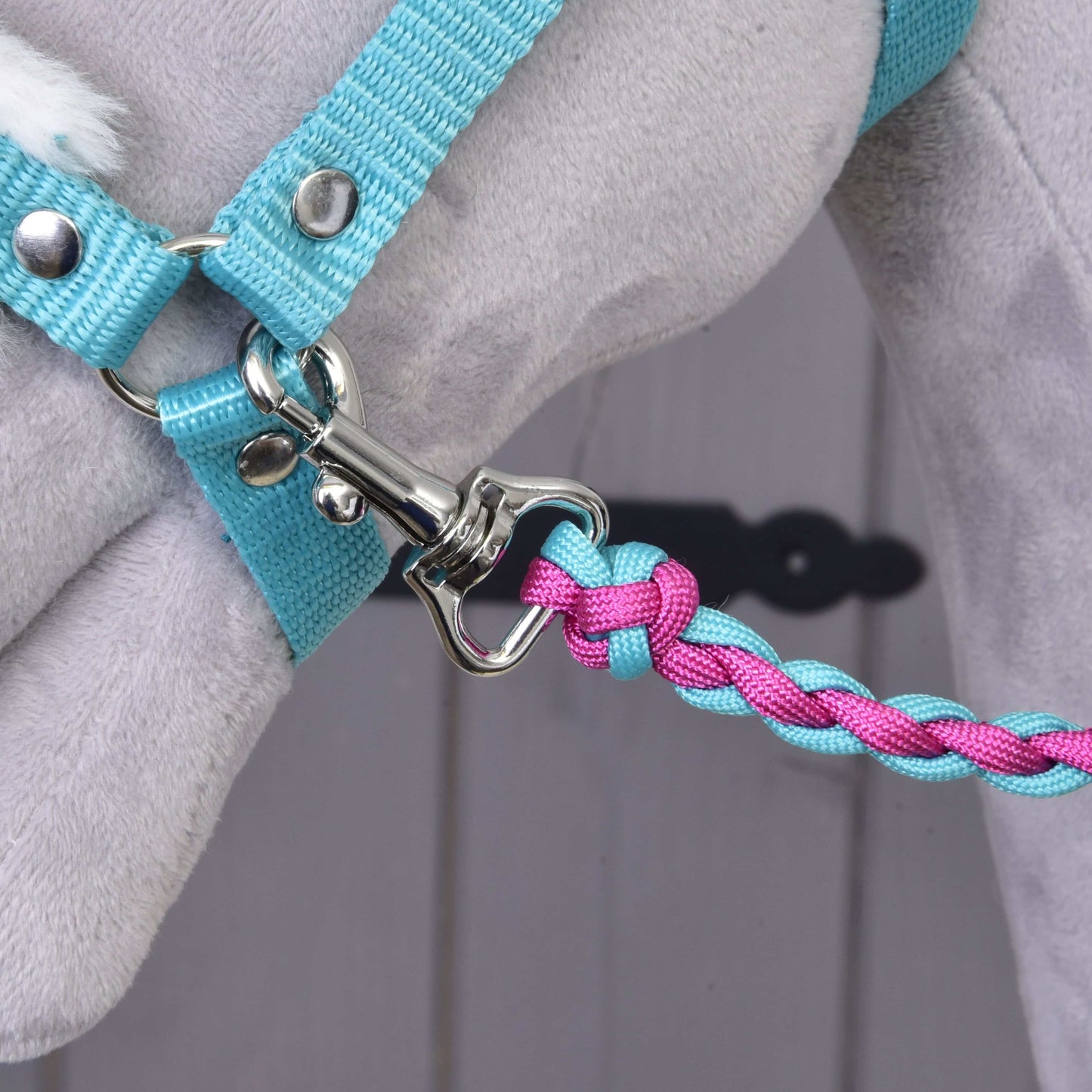 Detailní záběr na vodítko Set Perfect Combination Turquoise od Hobby Horse LarDen, pletené v modré a růžové barvě a připnuté ke světle modrému postroji na šedém plyšovém zvířeti – ideální pro doplňky pro hobby horse. Rozmazané pozadí.