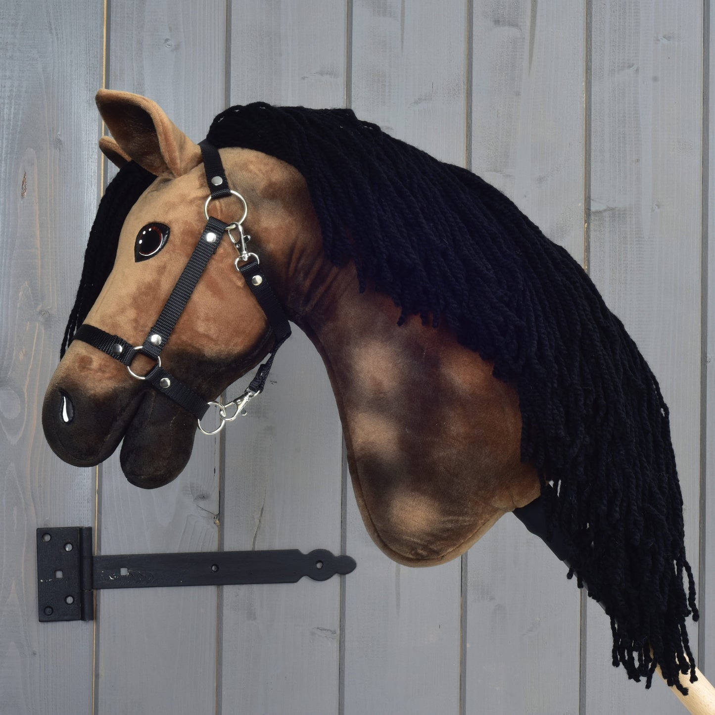 Plyšový hnědý Hobby Horse LarDen „Hobby Horse Stormy s černou ohlávkou“ s černou přízovou hřívou, nasazený na kovovém držáku u dřevěné stěny.