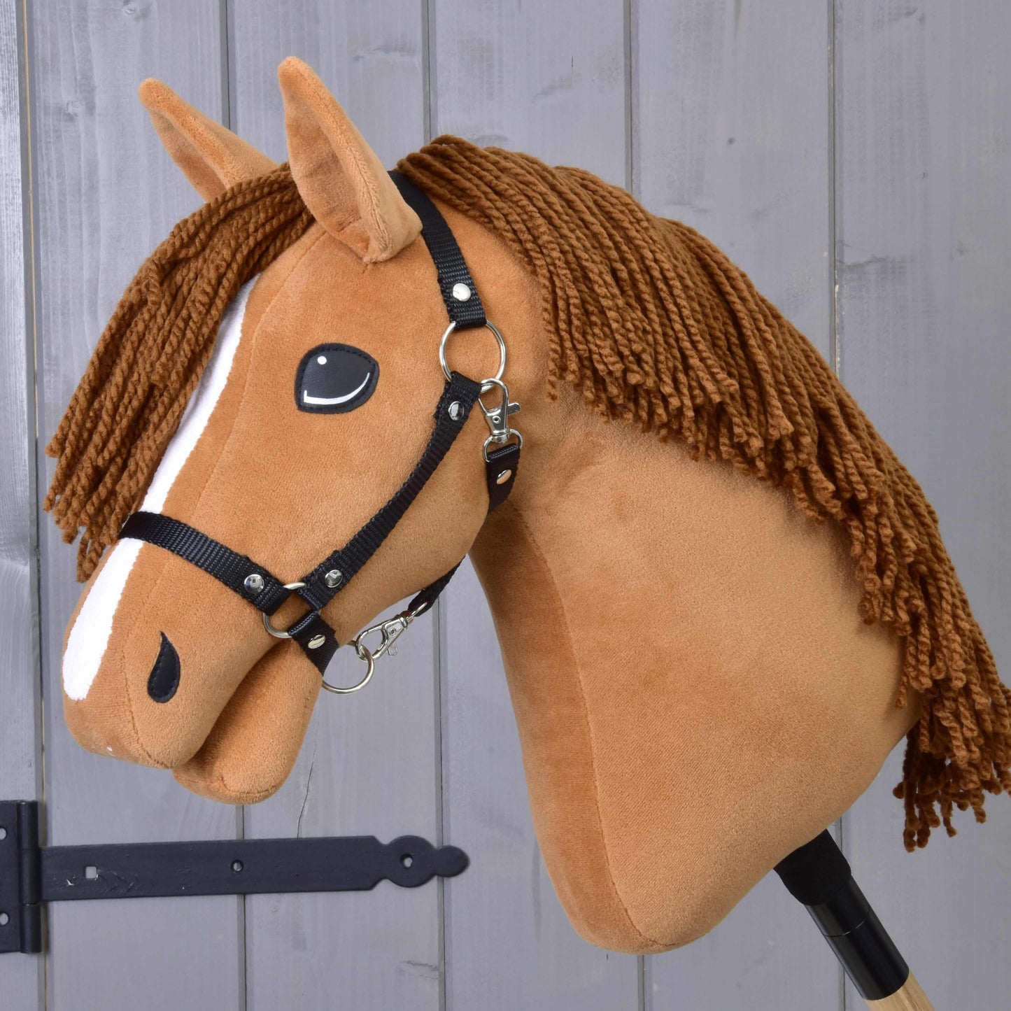 Hobby Horse Wendy s černou ohlávkou od Hobby Horse LarDen je plyšová hračka inspirovaná ryzkou klisnou s hnědou hřívou z příze, bílým pruhem na obličeji a černou ohlávkou – ideální pro nápaditou hru – zobrazená před dřevěným panelovým pozadím.