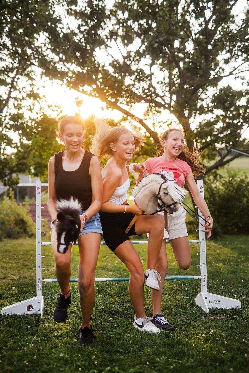 Tři mladé ženy se smějí, když venku běží a skáčou přes překážku, každá drží Hobby Horse, zatímco za nimi mezi stromy prosvítá slunce a ony si užívají zábavu při Hobby Horsing.