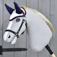 Čabraka Number 1 od Hobby Horse LarDen na ručně vyráběném šedém Hobby Horse – bílá s tmavě modrýma ušima pro ochranu proti hmyzu – s bílou hřívou, černou uzdečkou, nasazeném na dřevěné tyči před pozadím ze světlých dřevěných panelů.