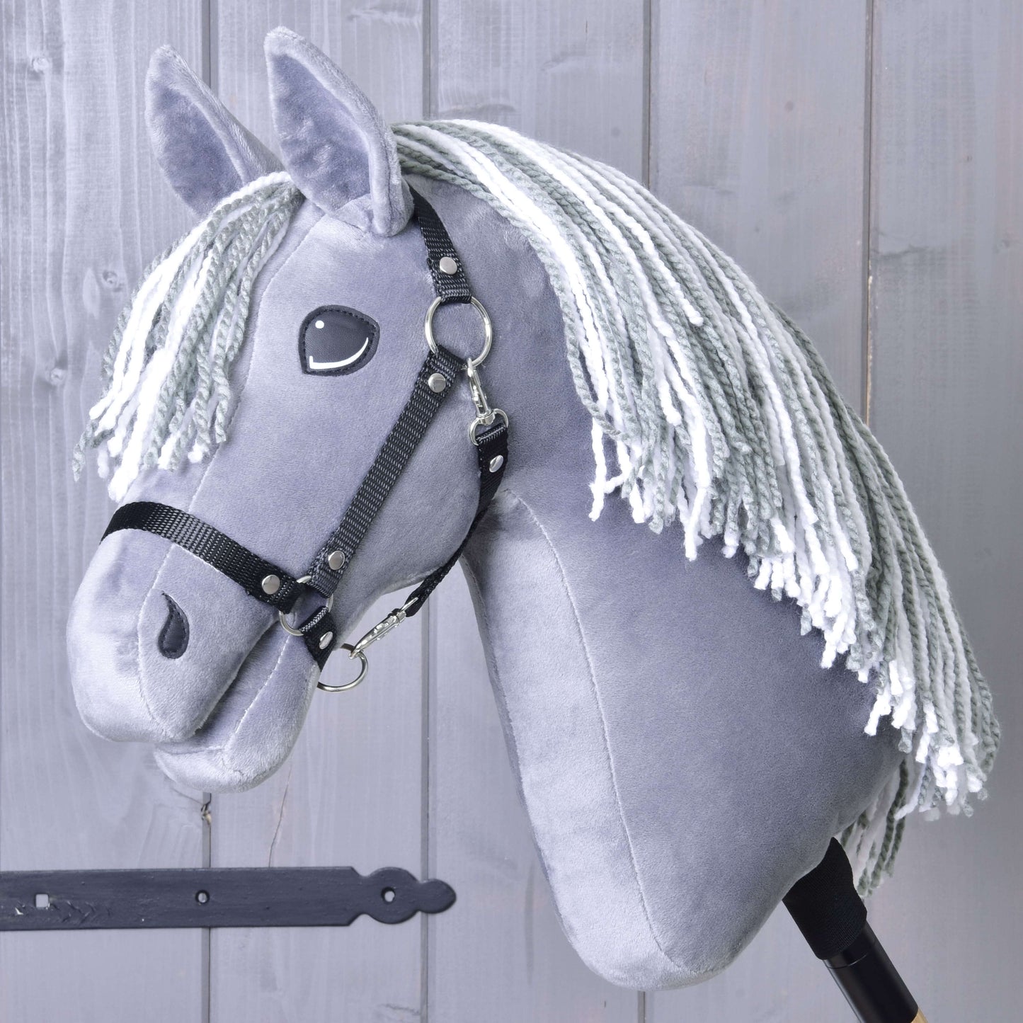 Hobby Horse Calypso s černou ohlávkou od Hobby Horse LarDen, inspirovaný holandským teplokrevníkem, má šedé zbarvení a bílou přízovou hřívu, je zobrazený na tyči před světle šedou dřevěnou stěnou.