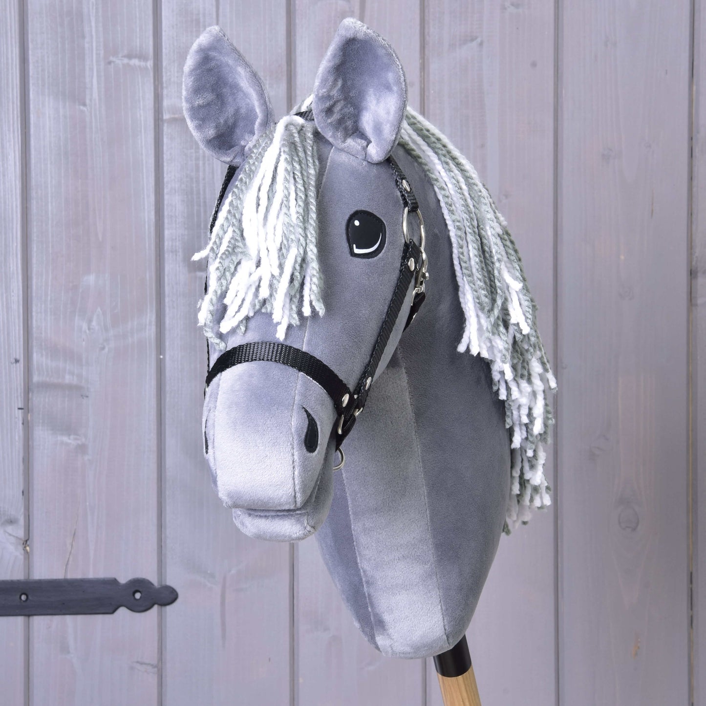 Hobby Horse Calypso s černou ohlávkou od Hobby Horse LarDen je šedý plyšový Hobby Horse inspirovaný holandským teplokrevníkem, s bílou přízovou hřívou a dřevěnou tyčí, zobrazený před šedou dřevěnou stěnou.