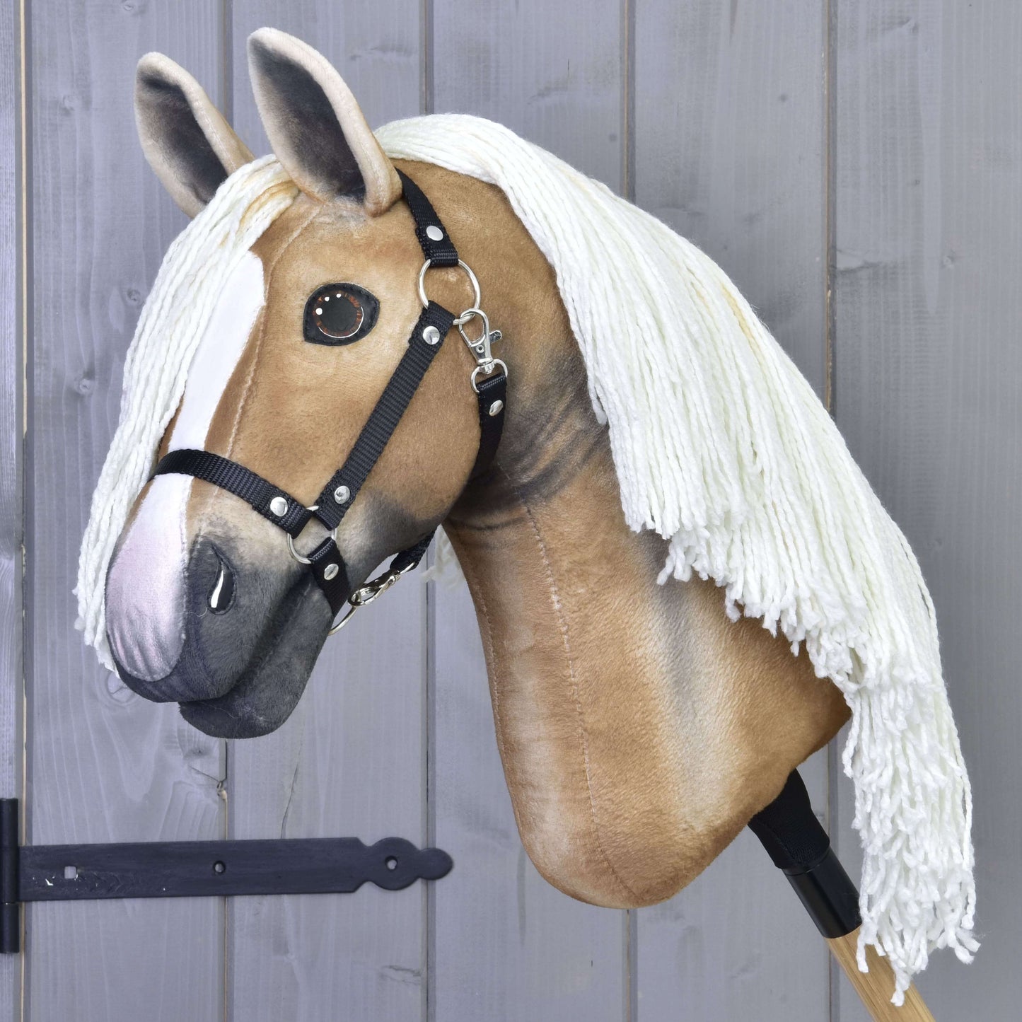 Hobby Horse Connie s černou ohlávkou od Hobby Horse LarDen s realistickým světle béžovým obličejem, bílou lysinou, černou ohlávkou a dlouhou bílou přízovou hřívou na dřevěné tyči, vystavený před šedou dřevěnou stěnou.