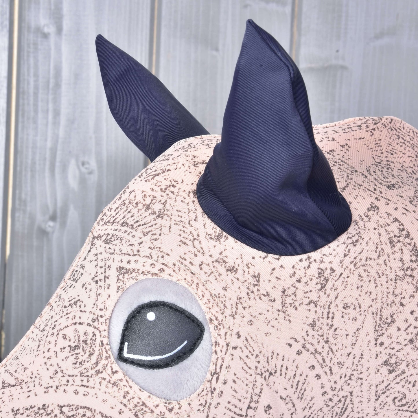 Detailní záběr na Hobby Horse ochranný oblek Cosmos (Lycra) od Hobby Horse LarDen s propracovanými vzory, velkým kulatým okem a dvěma špičatými tmavě modrými ušima na dřevěném pozadí.
