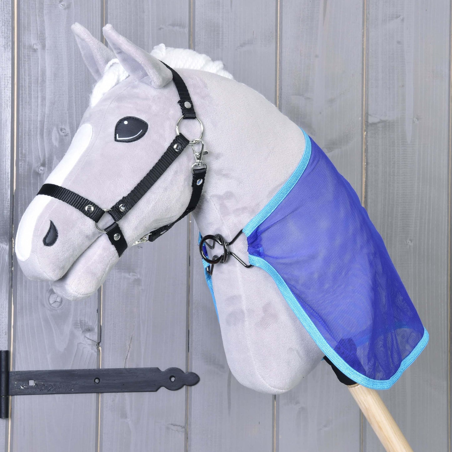 Šedá hračka Hobby Horse LarDen s černou uzdečkou a bílou hřívou má na sobě modrou Deka proti hmyzu. Kůň má oči ve stylu kreslené postavičky a je umístěný před dřevěnou stěnou.