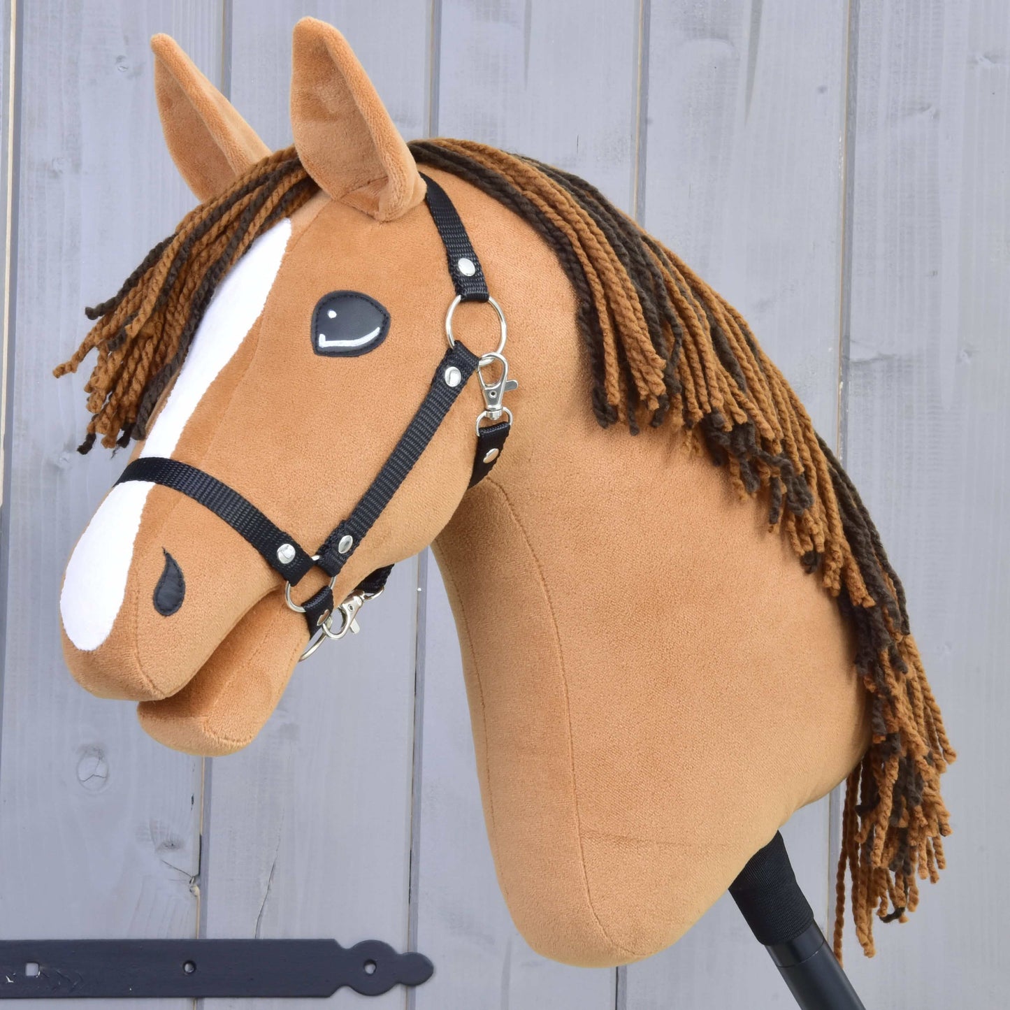 Ryzák hřebec Hobby Horse Edward-LD s černou ohlávkou od Hobby Horse LarDen má bílou lysinu a hnědou hřívu z příze, stojí na tyči před šedou dřevěnou stěnou.