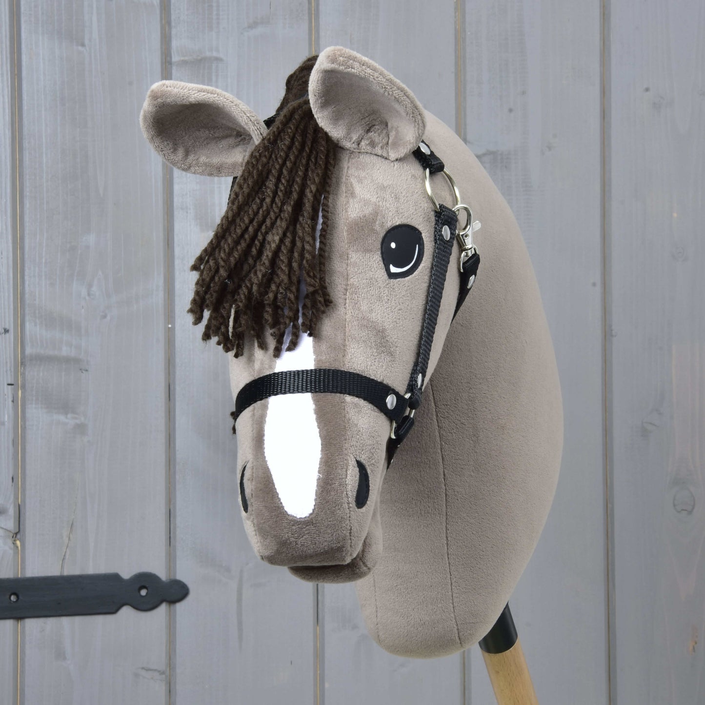 Hobby Horse Guccini s černou ohlávkou od Hobby Horse LarDen, s hnědou hřívou z příze, bílou lysinou a na dřevěné tyči, ideální pro jezdeckou hru, vystavený před šedou dřevěnou stěnou.